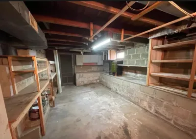 Unfinished basement with concrete block walls, water staining, exposed joists, and wood storage shelving.