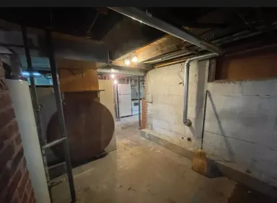 Unfinished basement with exposed pipes, a large rusted tank, brick walls, and a water heater.