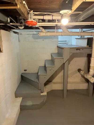 Unfinished basement stairwell with painted concrete walls, exposed pipes, and a bare bulb light fixture.
