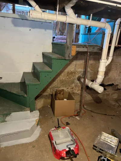 Basement stairwell mid-renovation with green-painted stairs, exposed pipes, and painting supplies on the floor.