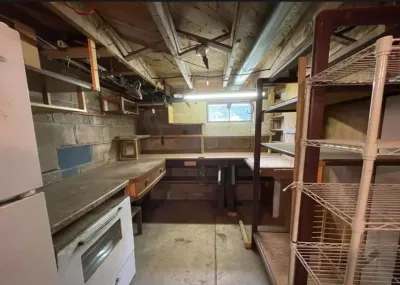 Unfinished basement workroom with exposed joists, concrete block walls, and cluttered shelving before renovation.