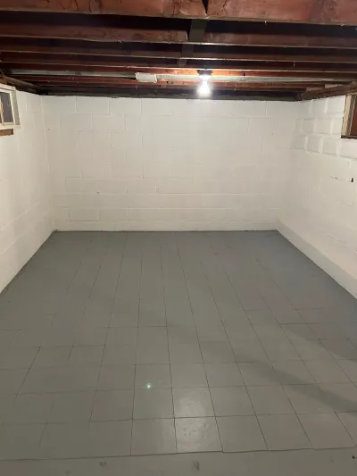 An empty unfinished basement with painted block walls, tile flooring, and exposed ceiling joists.