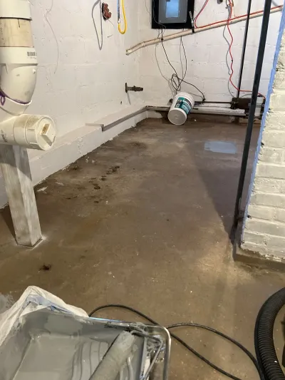 Basement under waterproofing repair with standing water, a support post, and a bucket of Drylok sealant.