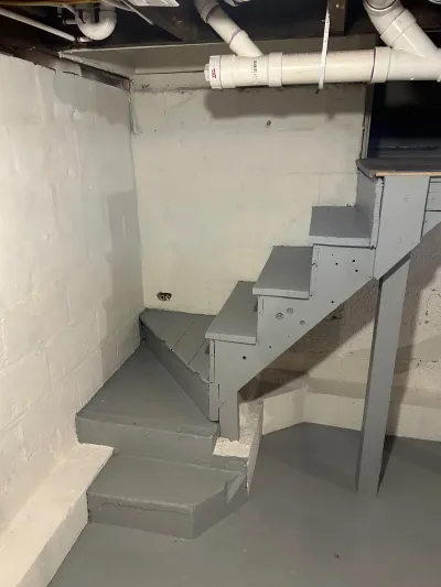 Basement stairwell with gray-painted wooden stairs, painted concrete block walls, and exposed overhead plumbing.