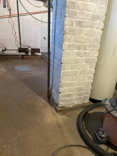 Basement corner with painted brick pier, exposed wiring, and a shop vacuum staged on a stained concrete floor.