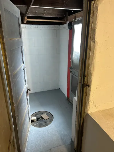 A basement utility room mid-renovation with painted block walls, a floor access hatch, and exposed ceiling joists.