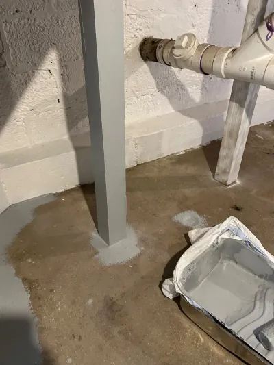 Basement floor mid-application of grey waterproof coating with paint tray and exposed plumbing pipes.