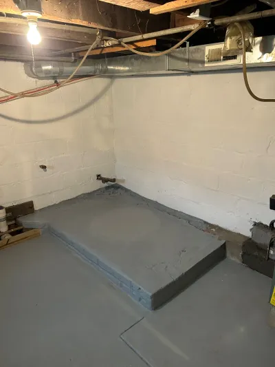 Unfinished basement with painted block walls, exposed joists and ductwork, and a raised concrete platform with a plumbing stu