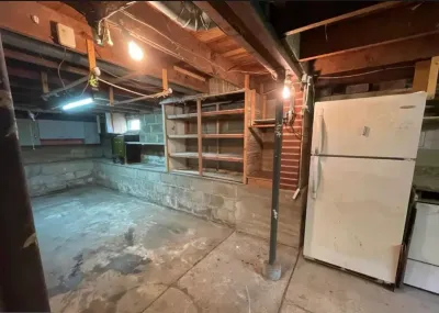Unfinished basement with exposed joists, concrete block walls, a support column, and an old refrigerator.