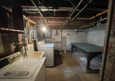 Unfinished basement with exposed joists, pipes, and wiring, showing a washer, dryer, and utility sink.