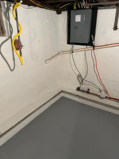Basement corner with painted block walls, exposed pipes and wiring, an electrical subpanel, and a drainage channel along the