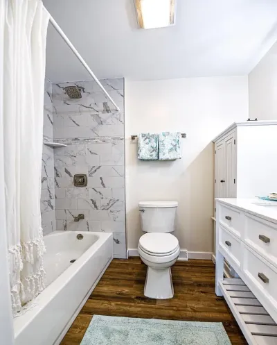 Finished bathroom with a marble-tile tub surround, wood-look flooring, and a white shaker vanity.