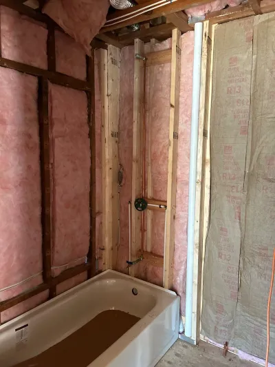 Bathroom under construction with exposed insulation, wood framing, plumbing rough-in, and a protected alcove tub.