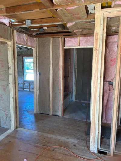 Interior under construction with exposed wood framing, pink insulation, and a bathroom rough-in visible through a doorway.