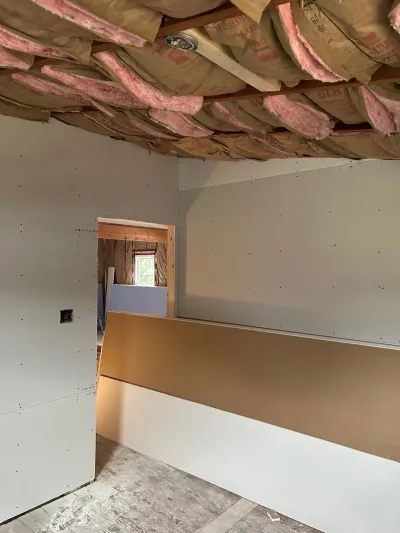 Room under construction with exposed ceiling insulation, hung drywall walls, and stacked drywall sheets.