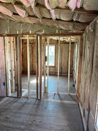 Interior under construction with exposed stud framing, pink batt insulation, and original hardwood floors.