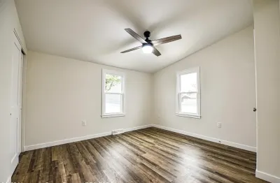 Empty bedroom with vaulted ceiling, dark plank flooring, and a ceiling fan between two windows.