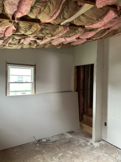 Room under construction with exposed ceiling insulation, unfinished drywall, and a stairway doorway visible.