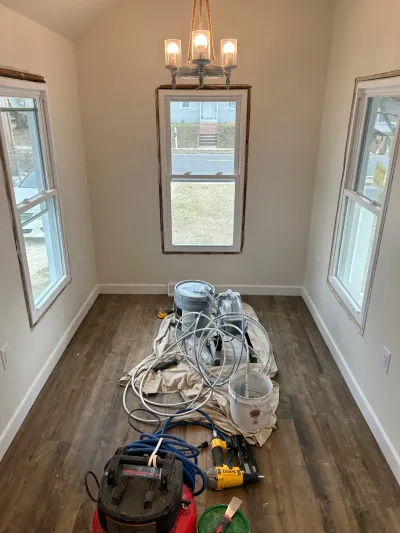 A sunroom mid-renovation with new flooring, untrimmed windows, and painting equipment scattered on a drop cloth.