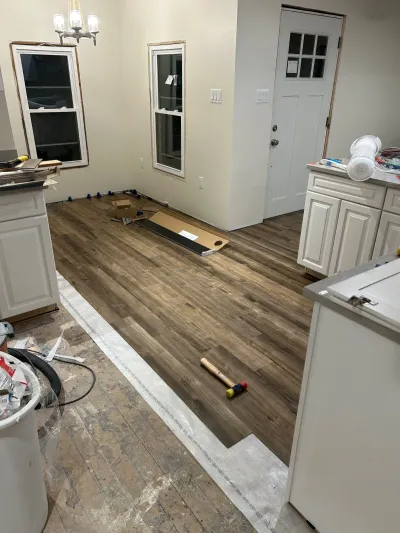 Kitchen mid-renovation showing new plank flooring being installed next to white cabinetry and exposed subfloor.