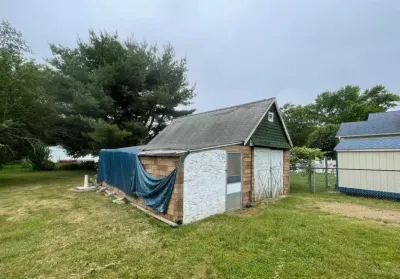 A weathered shed with concrete block walls, a tarp-covered section, and an aging shingle roof stands in an overgrown yard.