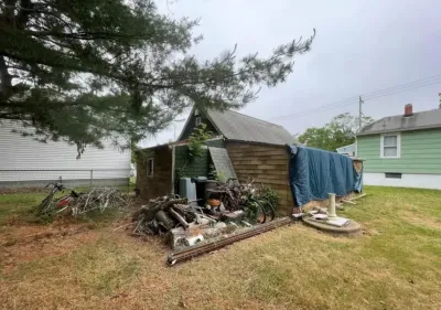 A weathered wood shed with a tarp-covered roof edge sits amid debris and firewood in a backyard.