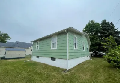 Side and rear view of a house with green siding, white trim, and a concrete block foundation on an overcast day.