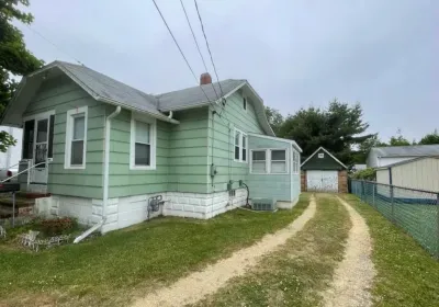 Side exterior of a green clapboard house with a gravel driveway leading to a small detached garage.