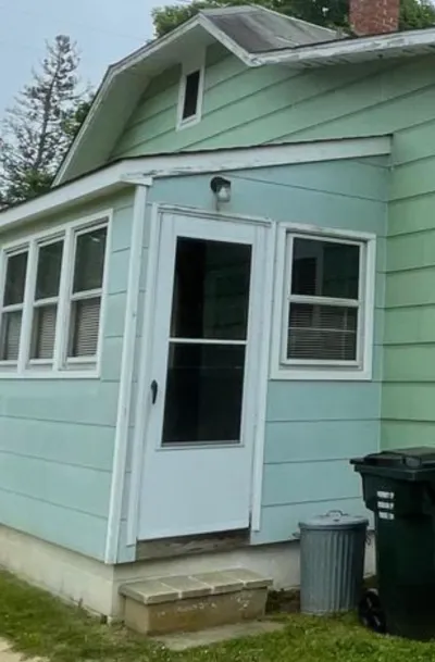 Side entrance of a house with faded green siding, white trim windows, and trash bins near the steps.