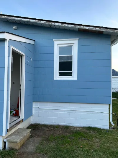 Side of a house with new blue siding installed, white trim, and an open door leading to an unfinished interior.