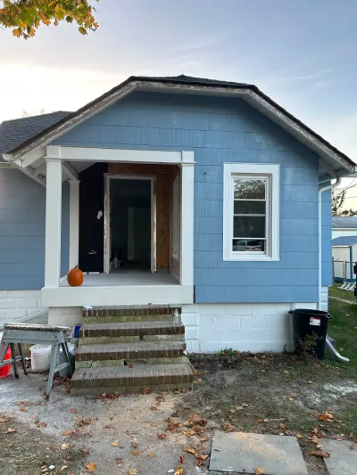 Front exterior of a house under renovation with new blue siding and an open, doorless entryway.