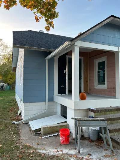 Bungalow exterior mid-paint job, with one wall finished in blue-grey siding and the rest still bare, porch and ladder visible