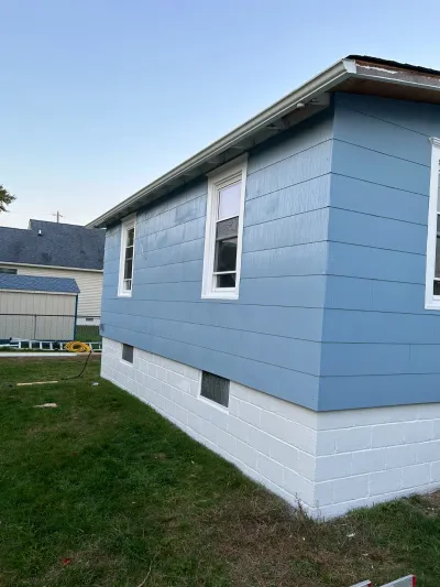 Corner of a house mid-renovation with new blue siding, white-painted foundation, and an unfinished roof eave.