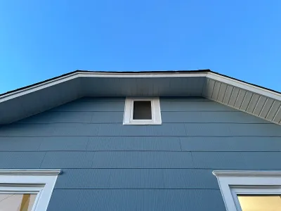 Looking up at a house's blue-grey gable end with white trim and a small window near the roof peak.