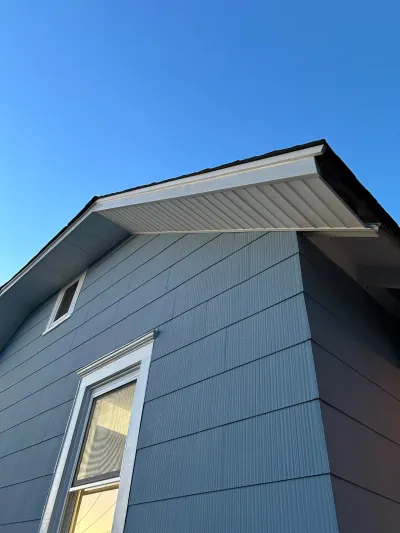 Upward view of a house's gable end showing blue-grey siding, white soffit and fascia, and two windows against a clear sky.