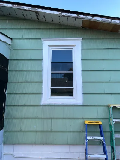 Exterior wall with a newly trimmed white window, worn eave paint, and ladders set up for ongoing repairs.