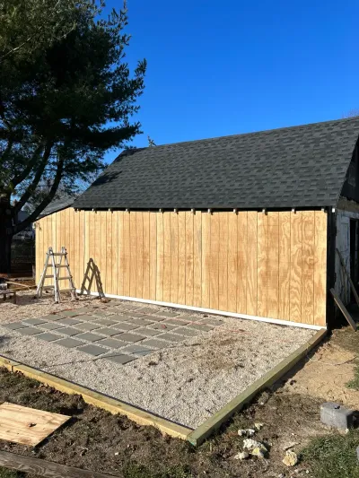 A barn under renovation with new plywood siding, a fresh shingle roof, and a paver patio being installed.