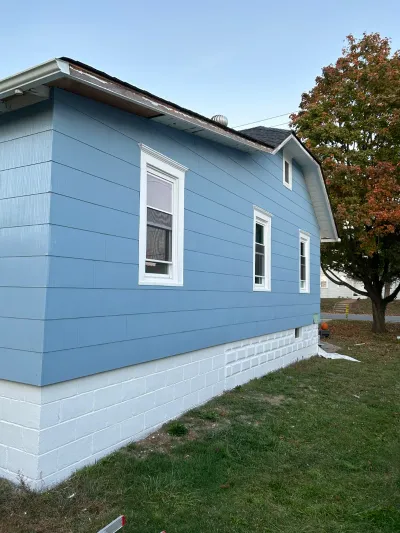 Side of a house with newly painted blue siding and white foundation, roof eaves still showing peeling paint.