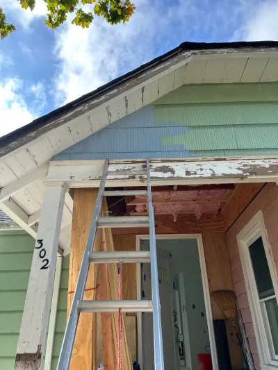 Home exterior mid-renovation with peeling paint, exposed insulation, and a ladder against the porch.