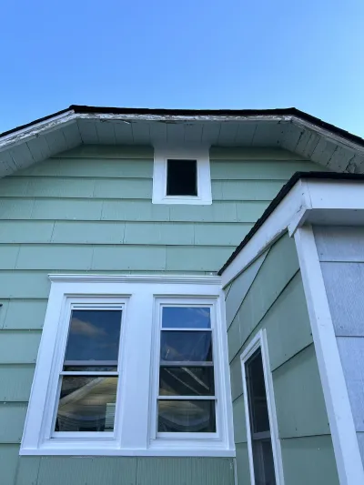 Weathered green house exterior showing peeling paint along the roofline above white-trimmed windows.
