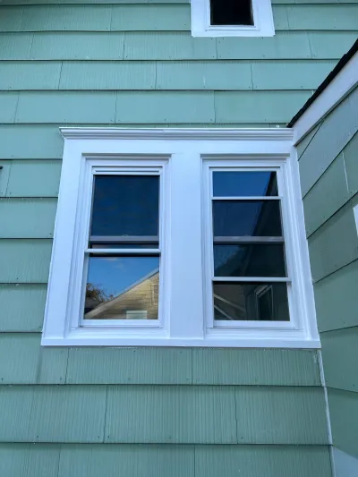 Two white-trimmed double-hung windows on a sage green clapboard house exterior.