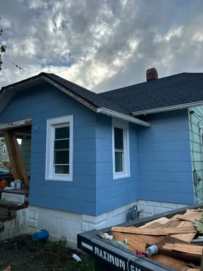 Partially renovated house exterior with new blue siding next to an area under demolition, dumpster in foreground.