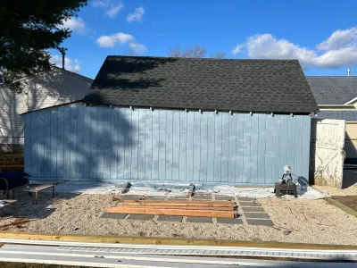 A shed with new blue siding and a shingled roof stands over a gravel yard with staged lumber and tarps.