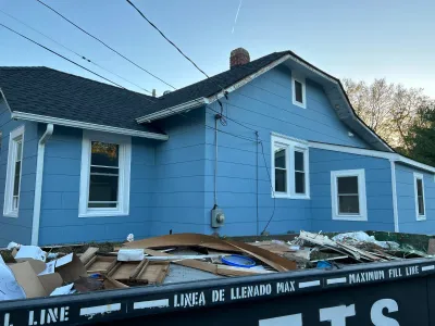 Blue house exterior with a debris-filled dumpster in front during a renovation project.
