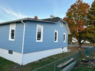 Side view of a house with new blue siding, white trim, and a painted block foundation, framed by a fall tree.