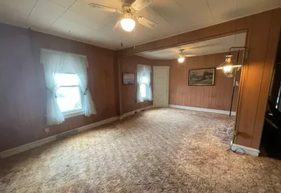 Empty living room with dated wood paneling, worn carpet, and a drop ceiling before renovation.