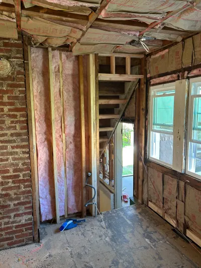 Gutted room mid-renovation with exposed brick, stud framing, insulation, and a view to a staircase and yard.