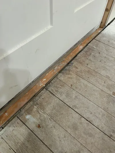 Close-up of unfinished subfloor and partially removed baseboard trim near a doorway during a renovation.