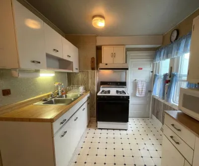 Dated galley kitchen with white laminate cabinets, gas range, and vinyl tile flooring before renovation.