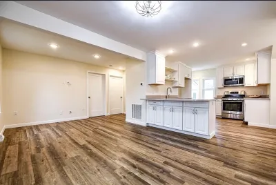 Open-concept white kitchen with a peninsula opening into an empty living area, both floored in wood-look plank.